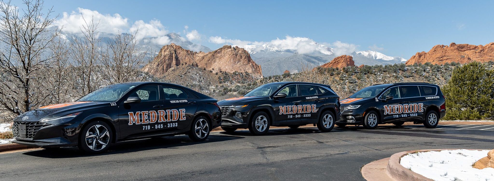 A lineup of MedRide Colorado's non-emergency medical transportation vehicles, including a sedan and SUVs, parked in front of the Garden of the Gods mountains.