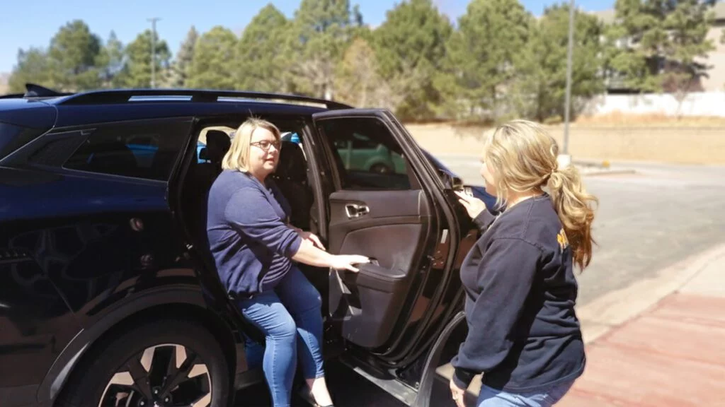 A MedRide Colorado driver assisting a female passenger exiting a black SUV for a medical appointment in a sunny outdoor setting.